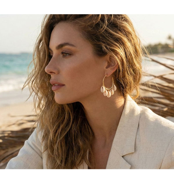 Woman on a beach with ocean view wearing a beige jacket and stunning cowrie hoop earrings.