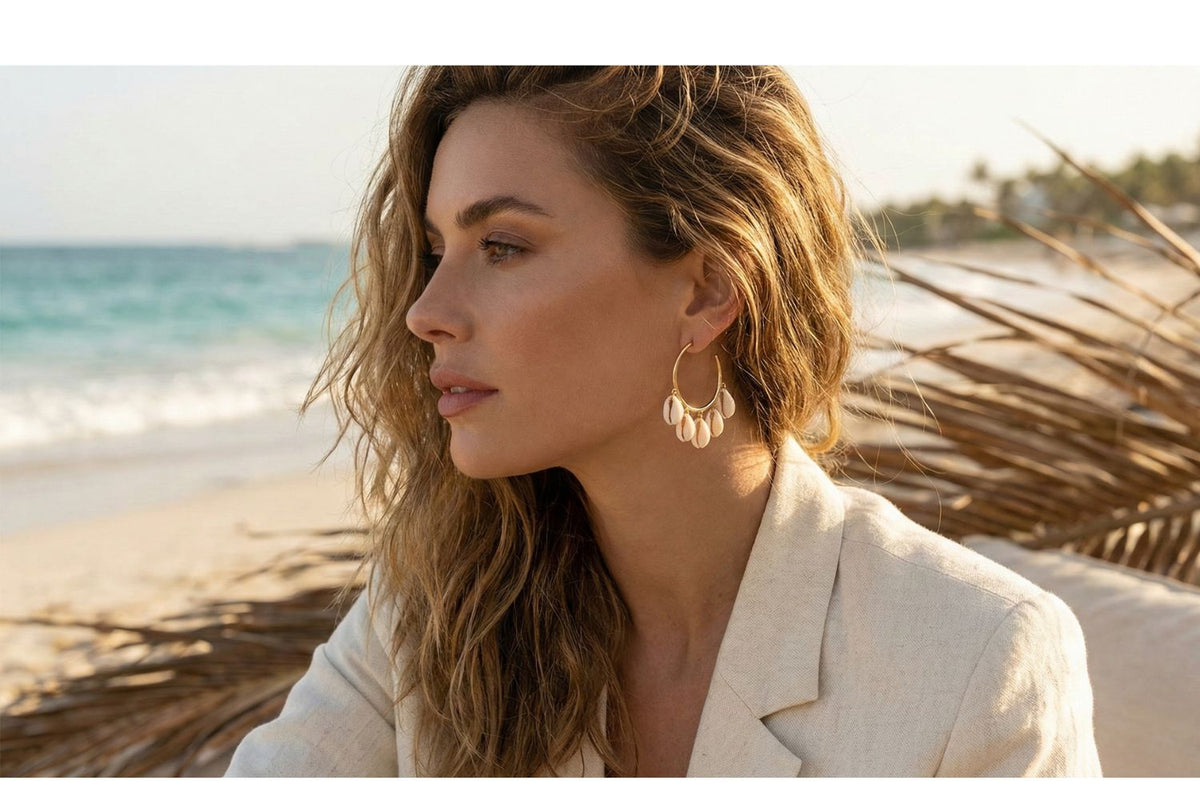 Woman on a beach with ocean view wearing a beige jacket and stunning cowrie hoop earrings.