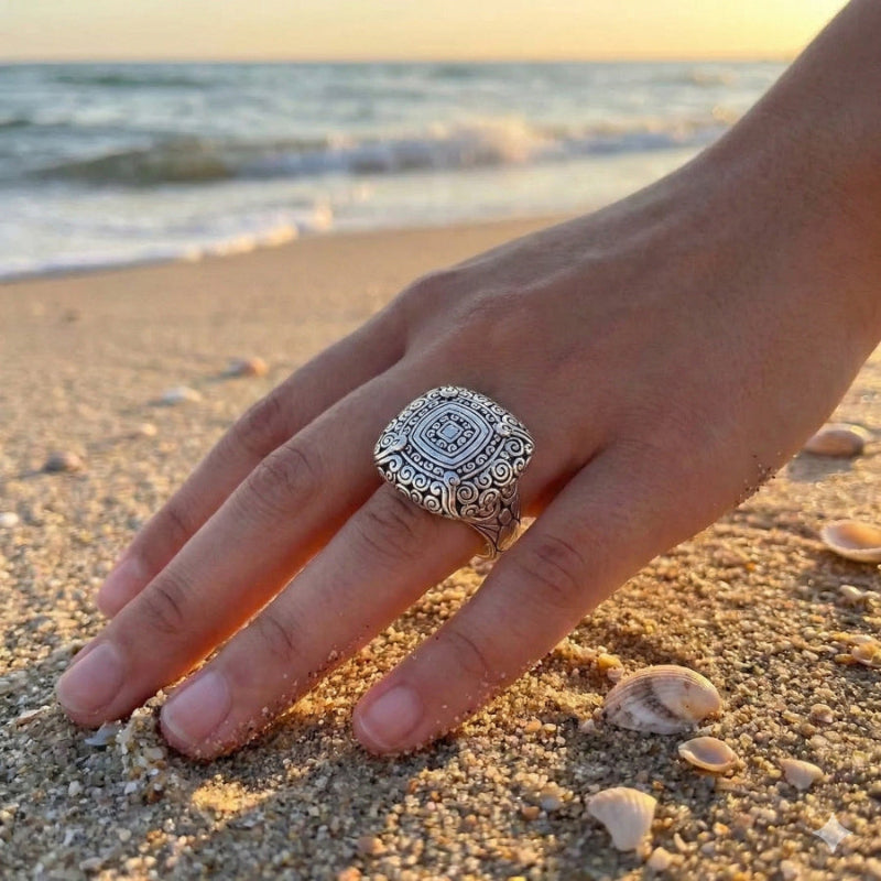 Hand wearing a silver ring on a sandy beach with shells and ocean waves in the background