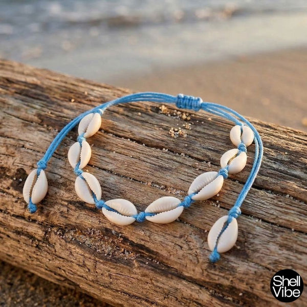 Cowrie shell anklet with blue cord on a wooden log by the beach, 'Shell Vibe' brand visible.