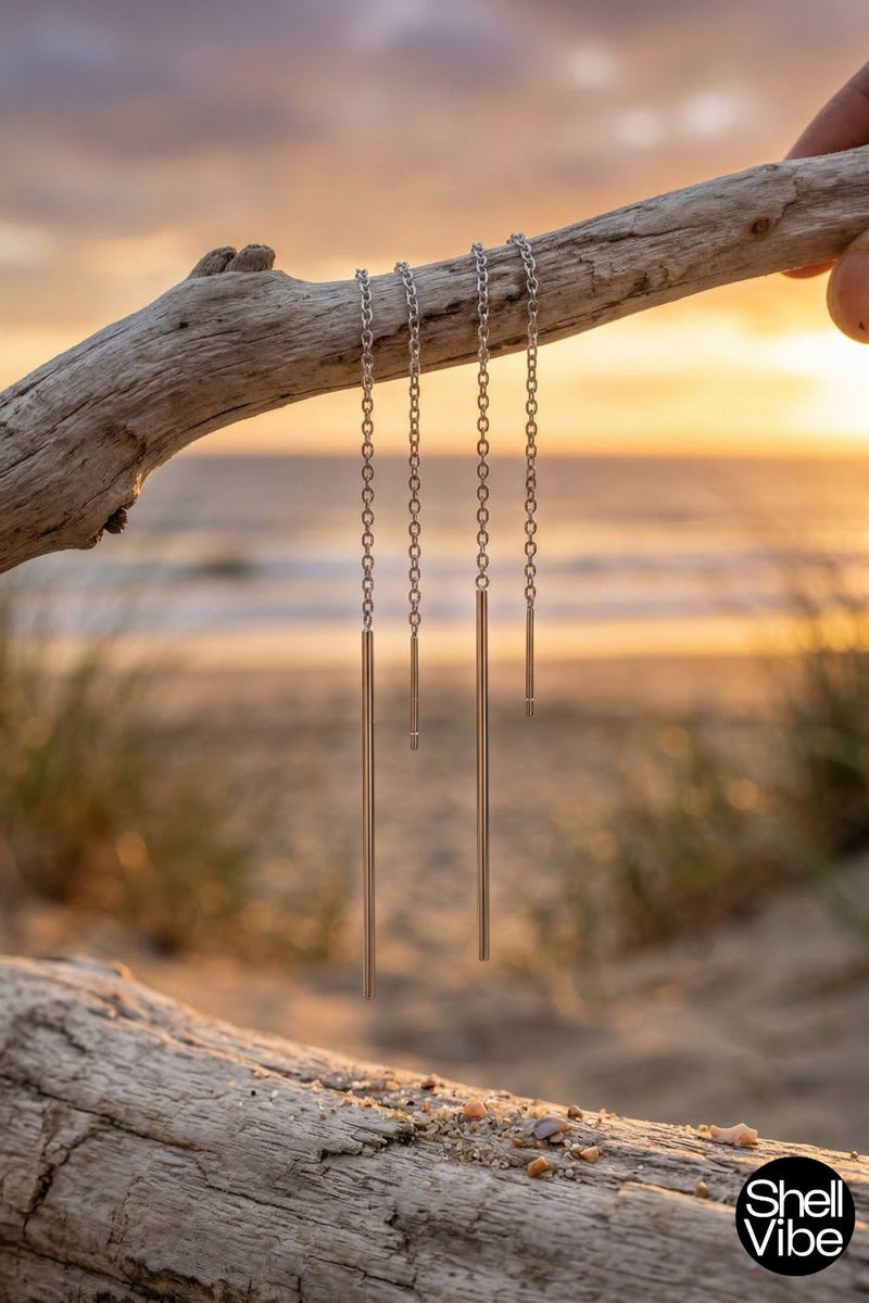 Two sterling silver earrings hanging from a branch on a beach with a sunset in the background.