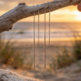 Two sterling silver earrings hanging from a branch on a beach with a sunset in the background.