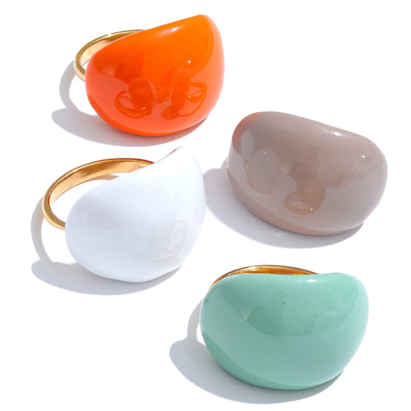 “Gold-plated enamel dome rings in orange, white, beige and mint displayed beside a large seashell on a textured stone surface.”
