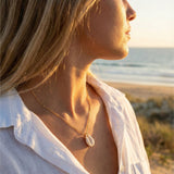 Woman in a white shirt standing  AND WEARING A COWRIE SHELL NECLACE on a beach with sunlight casting a warm glow.