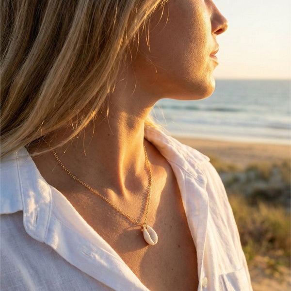 Woman in a white shirt standing  AND WEARING A COWRIE SHELL NECLACE on a beach with sunlight casting a warm glow.