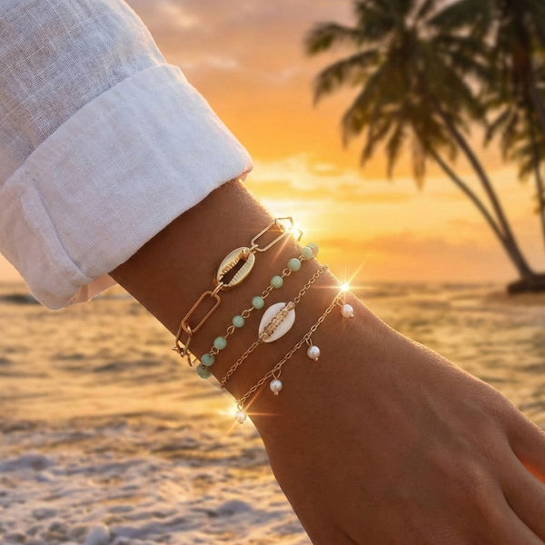 Hand wearing layered shell and gold chain bracelets with a sunset and palm tree in the background
