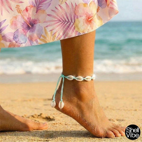 Person wearing a cowrie shell anklet on a beach with floral skirt and ocean in background