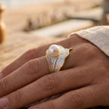 Hand wearing a sterling silver and gold ring with a pearl on a wooden surface, blurred background