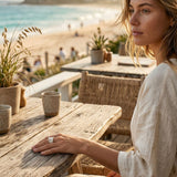 Woman sitting at a wooden table by the beach with a scenic view, wearing a sterling silver and gold pearl ring with a blurred beach background