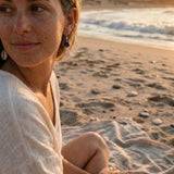 Woman sitting on a beach with ocean waves and sunset in the background, wearing cowrie earring dangles