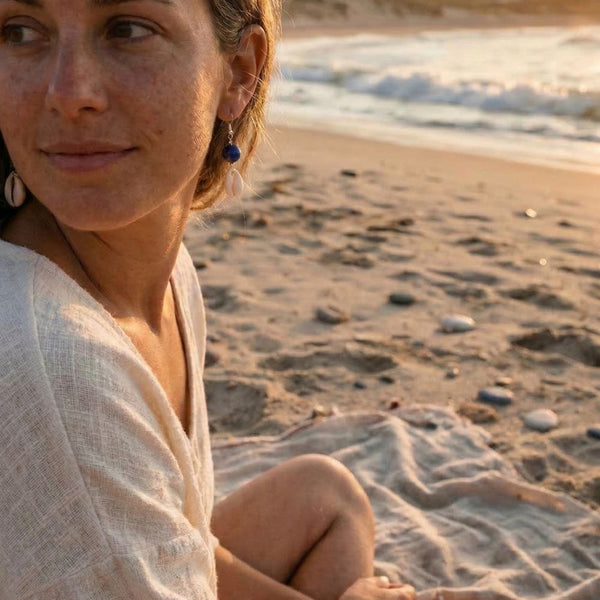Woman sitting on a beach with ocean waves and sunset in the background, wearing cowrie earring dangles