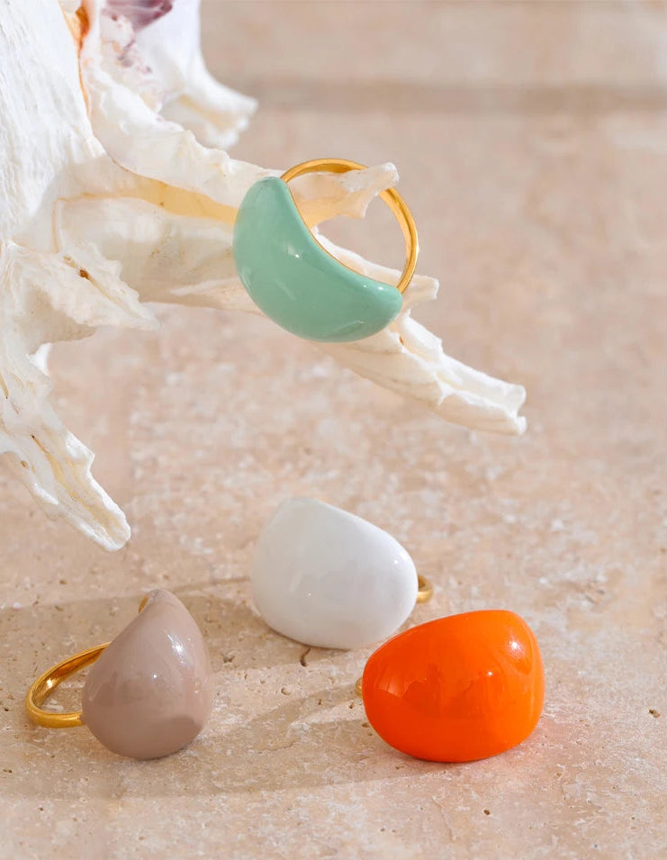 “Mint enamel dome ring resting on a seashell with additional rings in white, beige and orange displayed below on a textured stone background.”
