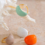 “Mint enamel dome ring resting on a seashell with additional rings in white, beige and orange displayed below on a textured stone background.”
