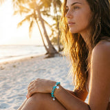 Woman sitting on a beach with palm trees and sunset in the background, wearing a turquoise bracelet.