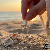 Sterling silver starfish-shaped pendant held by a hand on a sandy beach with sunset in the background