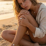 Woman sitting on a beach with a scenic background, wearing a light-colored shirt and mother of pearl blossom jewelry.