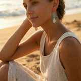 Woman sitting on a beach wearing COWRIE earrings with a blurred background