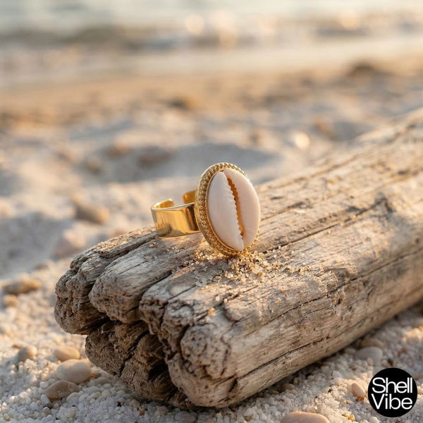Gold ring with a cowrie shell on a piece of driftwood on a beach, featuring the 'Shell Vibe' brand.