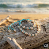 Blue cord cowrie shell necklace and  bracelet on a piece of driftwood with ocean in the background
