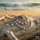 Brown cord cowrie shell necklace and  bracelet on a piece of driftwood with ocean in the background