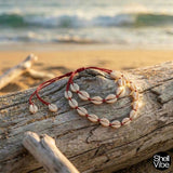 Red cord cowrie shell necklace and  bracelet on a piece of driftwood with ocean in the background