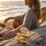 Woman sitting on sand by the ocean with a golden cowrie ring on driftwood