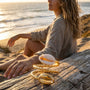 Woman sitting on sand by the ocean with a golden cowrie ring on driftwood
