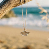 Shell-shaped pendant on a chain hanging from a branch with a beach background