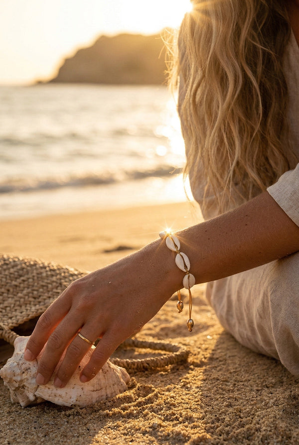 Woman sitting on a beach at sunset, wearing a cowrie shell bracelet.