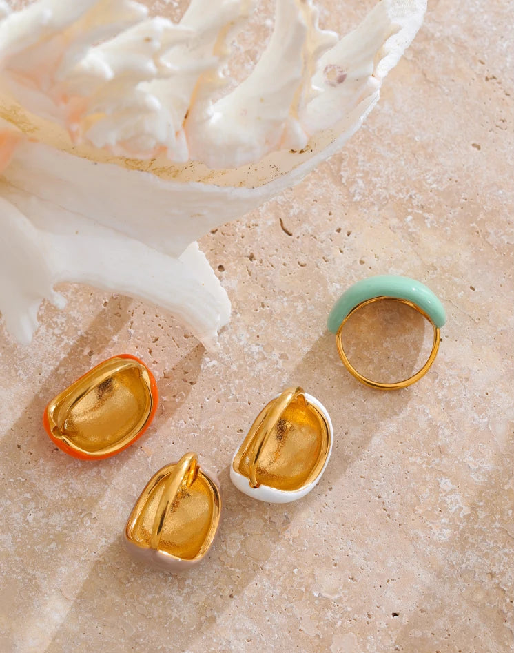 Close-up of four gold-plated enamel dome rings in vibrant colors—orange, mint, beige and white—arranged on a white background.”
