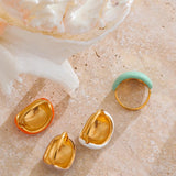 Close-up of four gold-plated enamel dome rings in vibrant colors—orange, mint, beige and white—arranged on a white background.”
