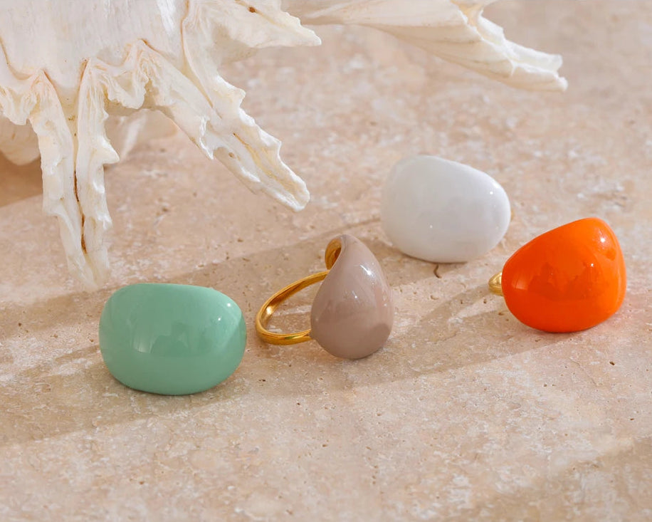“Set of colorful enamel dome rings arranged on a stone surface near a white seashell, showcasing orange, mint, beige and white tones.”
