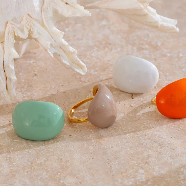 “Set of colorful enamel dome rings arranged on a stone surface near a white seashell, showcasing orange, mint, beige and white tones.”
