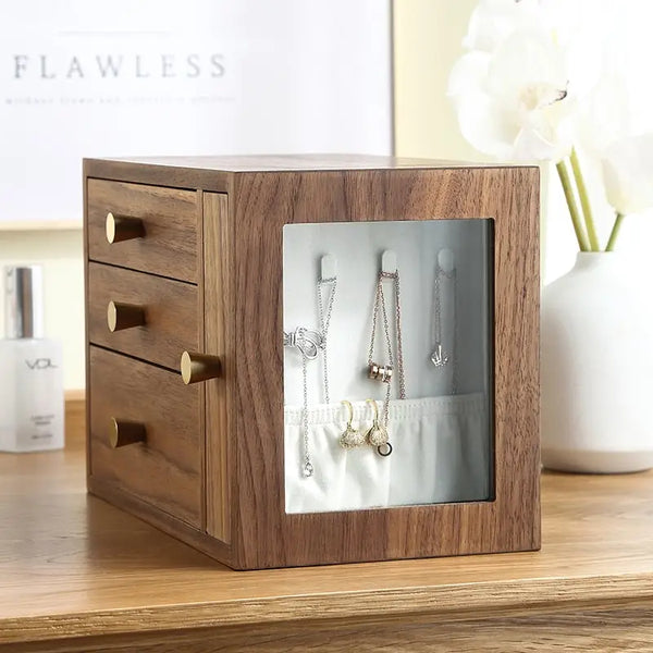 Walnut wooden jewelry box with a glass side door displaying hanging necklaces and earrings, placed on a minimalist vanity table with soft decor.

